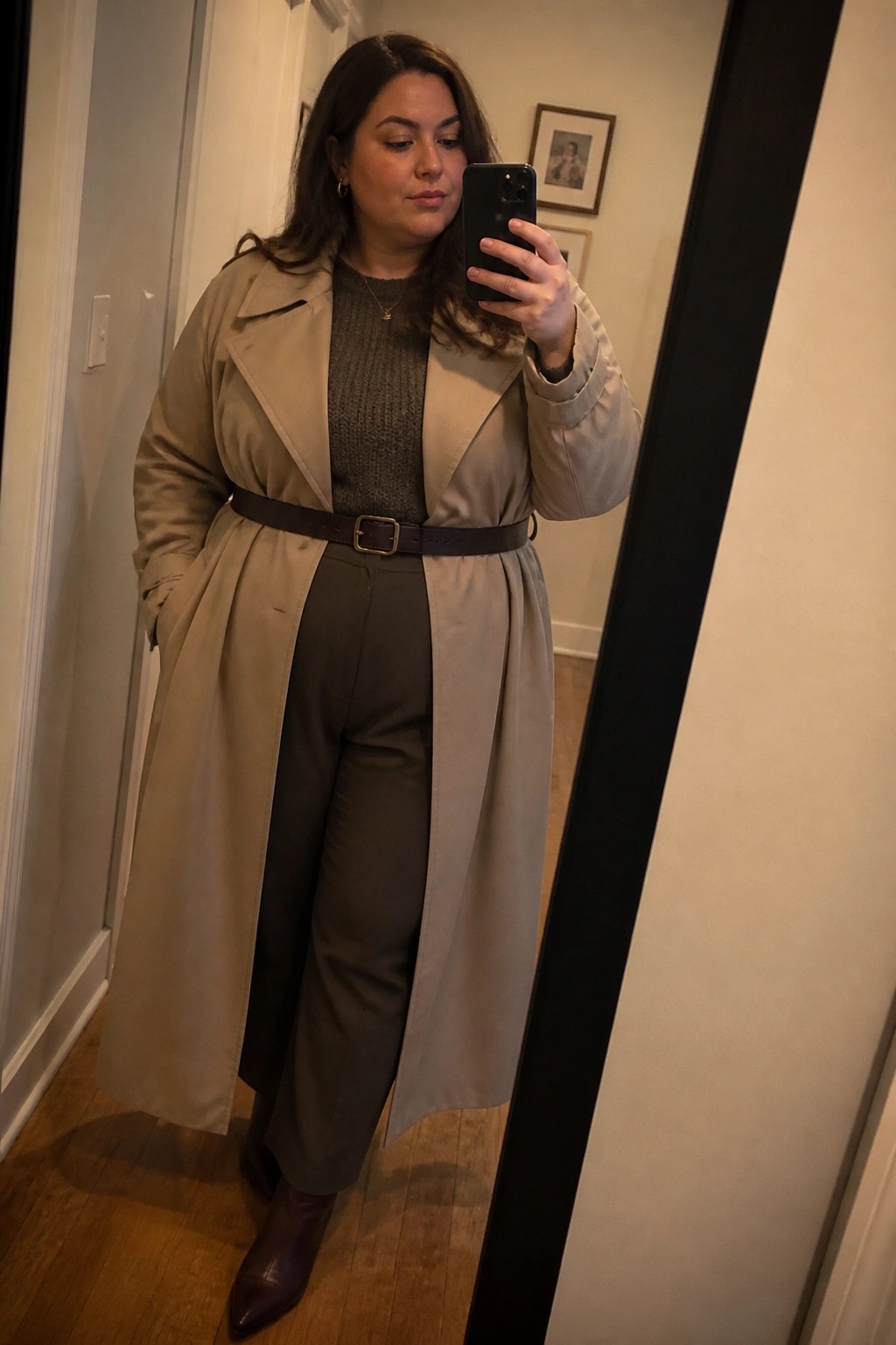 Woman in beige trench coat taking mirror selfie in hallway with phone