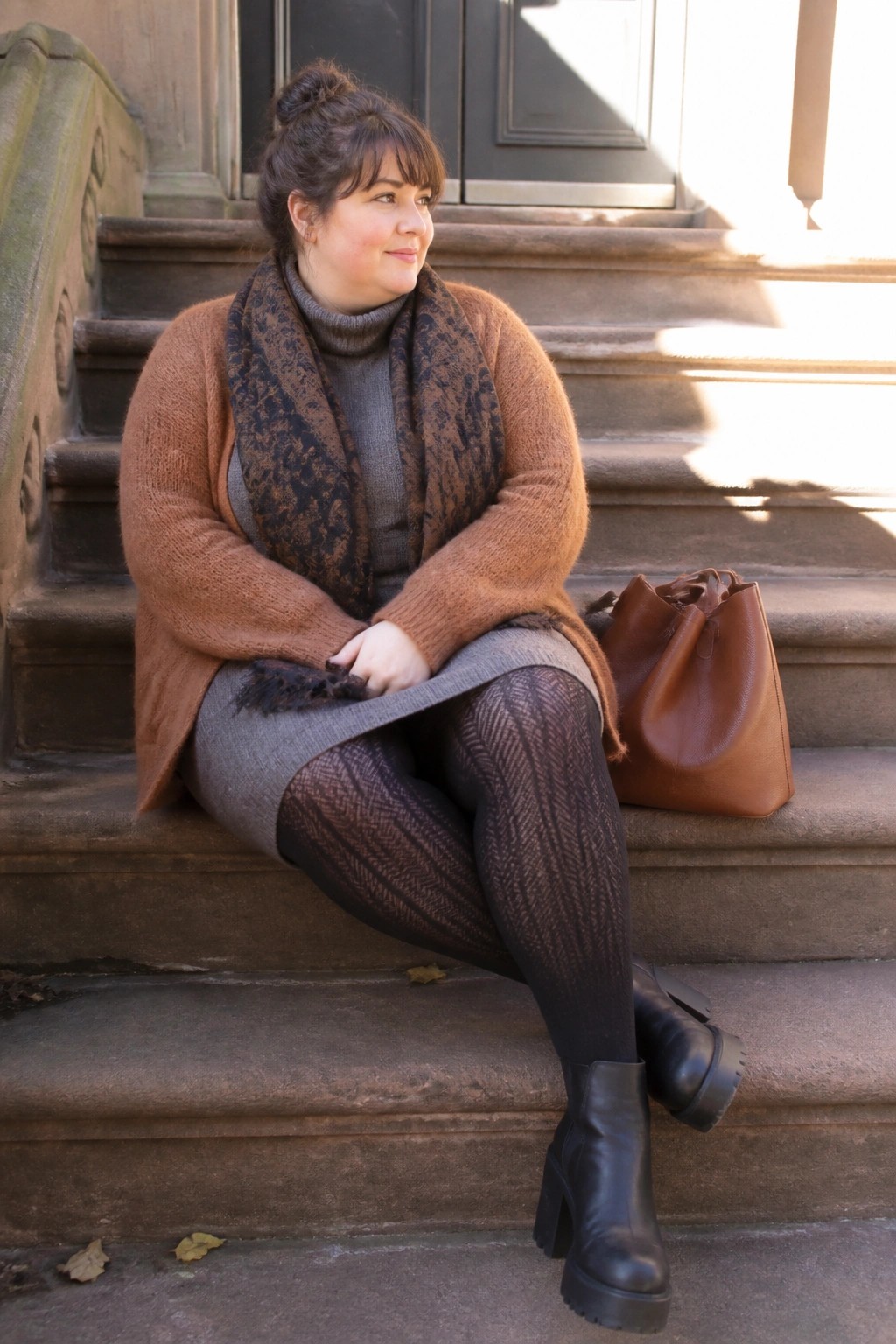 Woman in brown cardigan and scarf sits on stone steps with leather bag