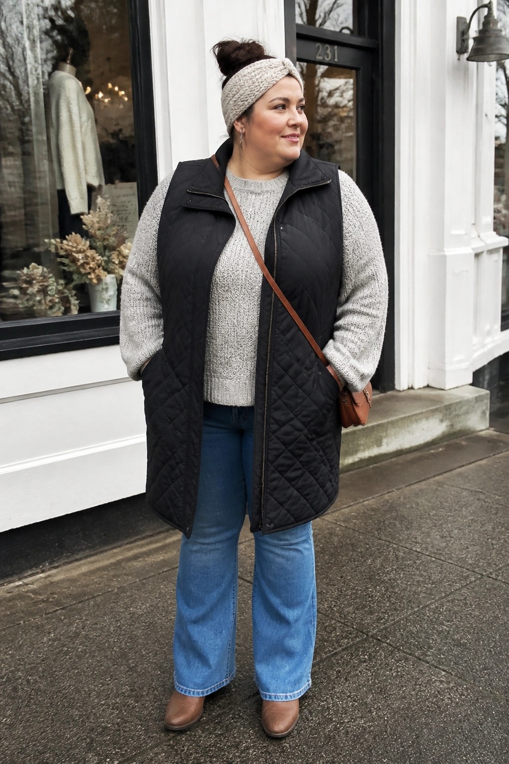 Plus size woman wearing a black quilted puffer vest over a gray marled knit sweater paired with blue wide leg jeans, brown ankle boots, a beige knit headband, and a brown crossbody bag.