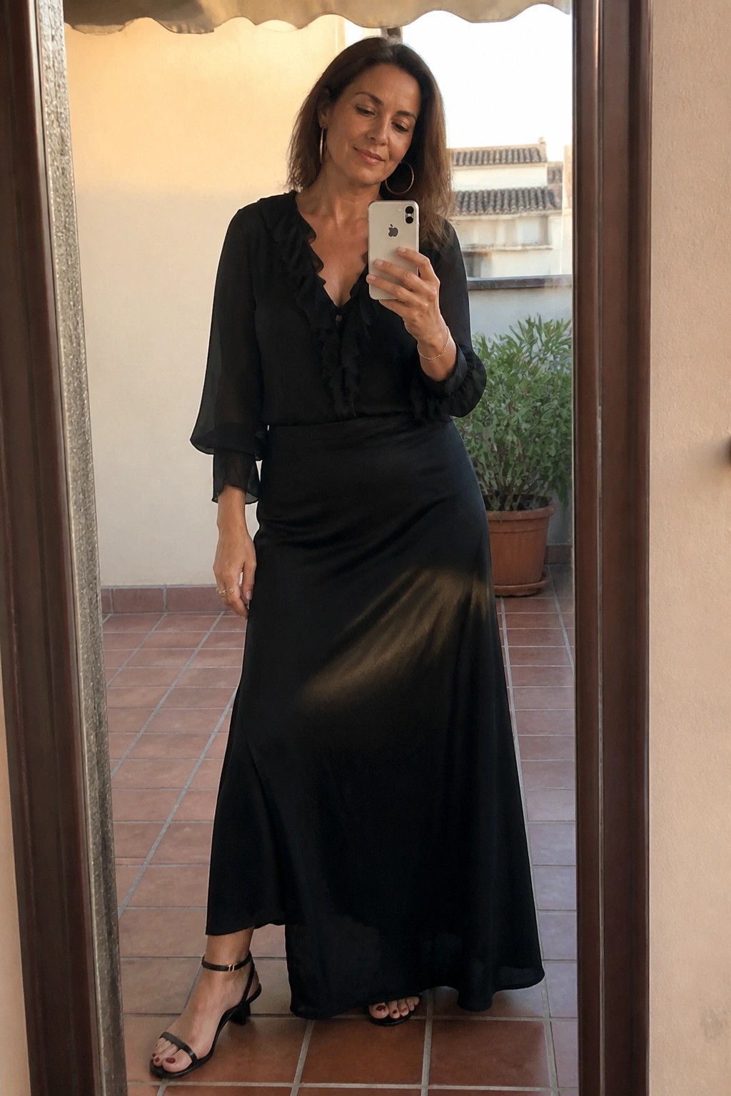 Woman in black ruffled blouse and long satin skirt taking mirror selfie with iPhone.