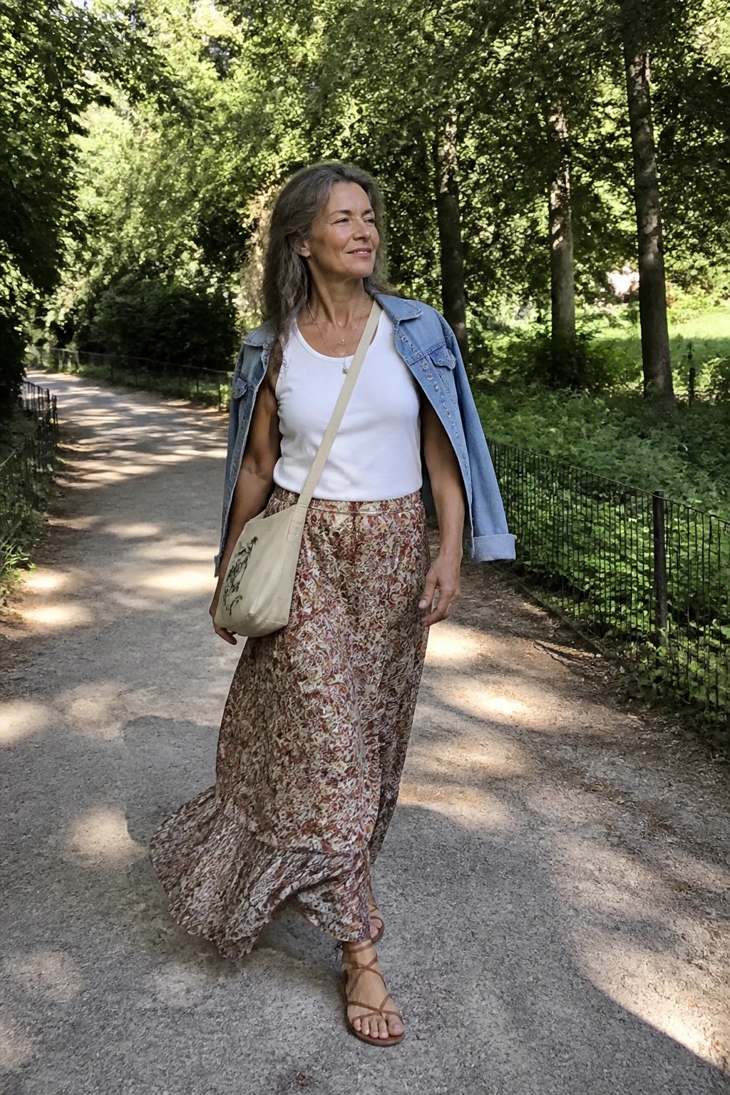 Woman over 50 wearing a white tank top, light denim jacket draped over shoulders, floral maxi skirt, beige crossbody bag, layered necklaces and strappy sandals on a park path.