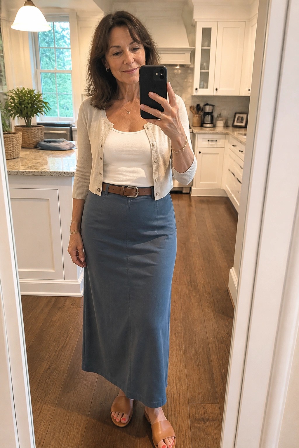 Woman over 50 in mirror selfie wearing blue denim maxi skirt, white tank top, beige cardigan, brown belt, and brown slide sandals with gold jewelry.