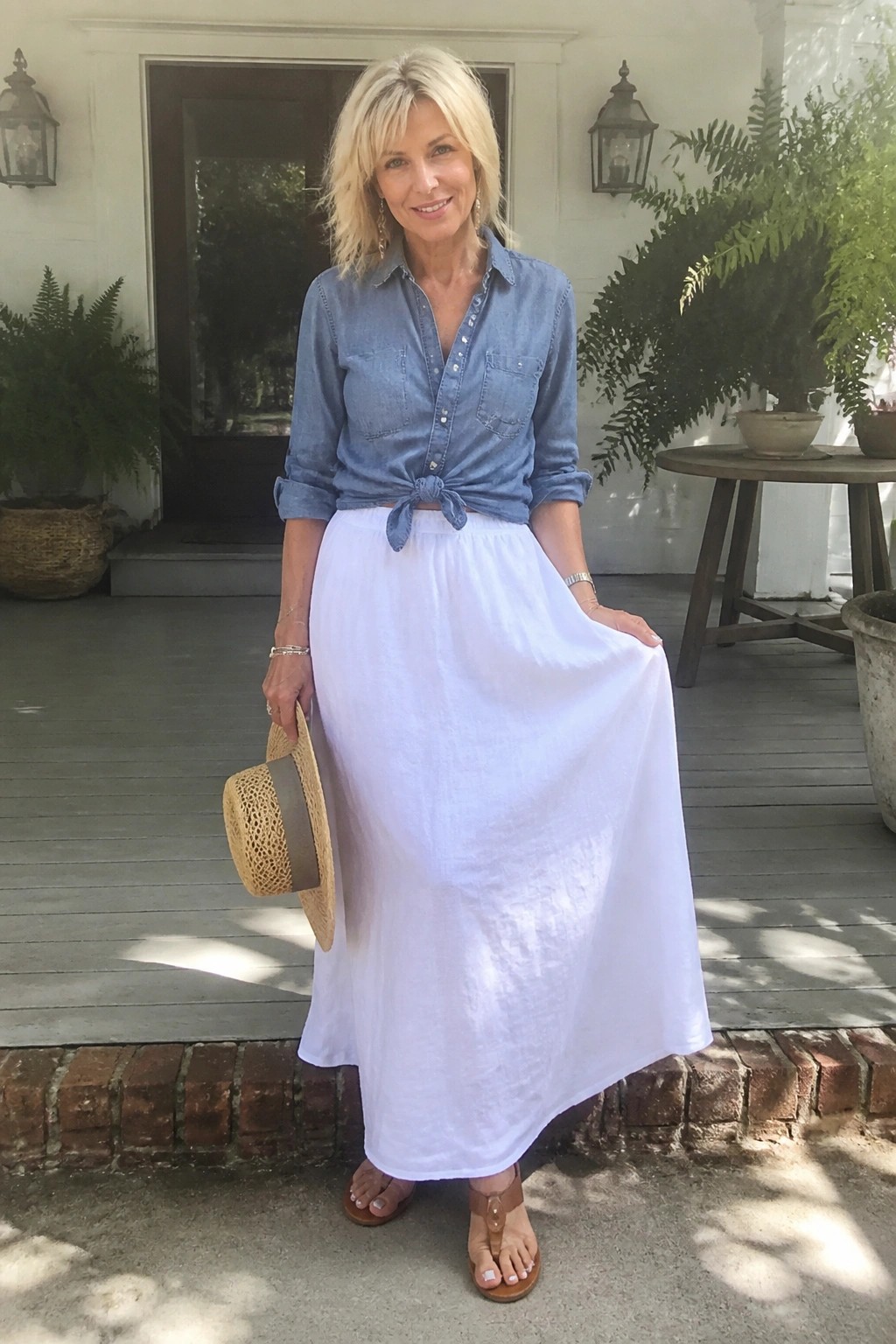 Blonde woman in knotted blue denim shirt and flowing white skirt holding straw hat.