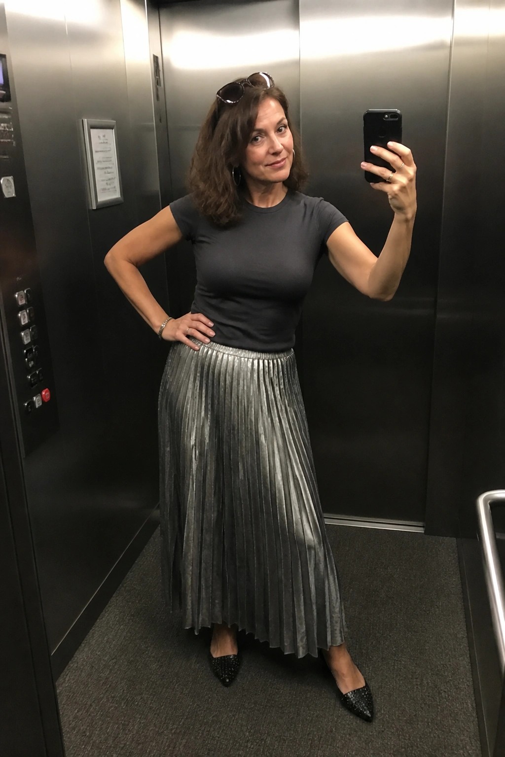 Woman in gray top and metallic silver pleated skirt taking selfie in elevator.