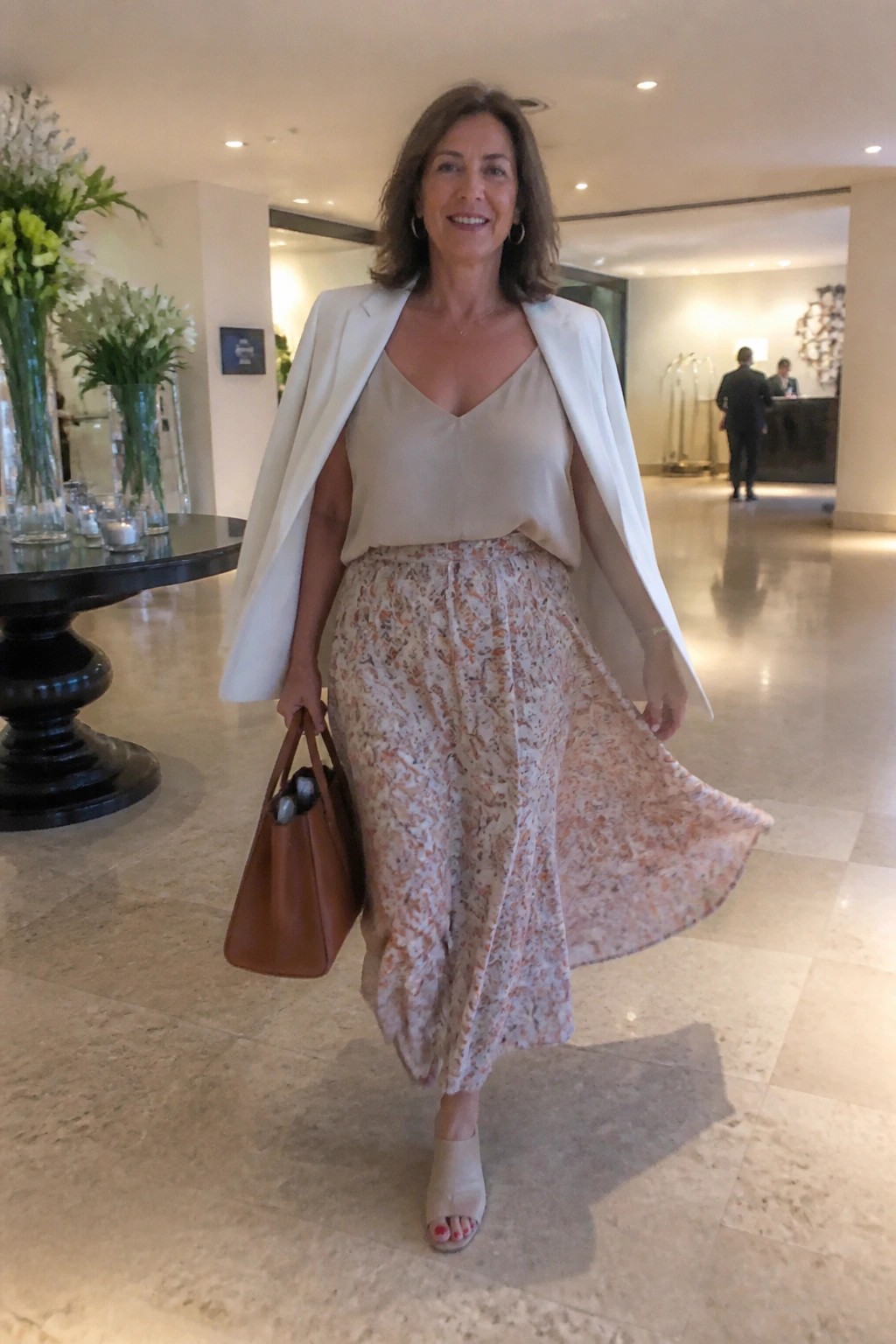 Woman over 50 in a white blazer draped over shoulders with beige camisole and floral maxi skirt in beige and orange tones paired with brown handbag and neutral heels.