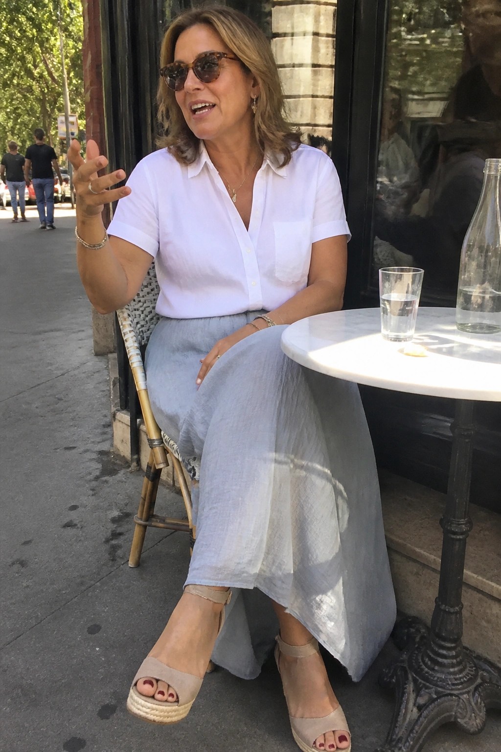 Woman in white shirt and blue skirt gesturing at outdoor cafe table