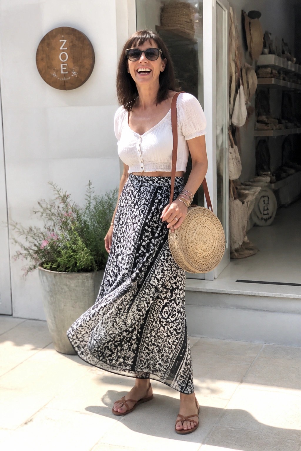Woman over 50 in a white short-sleeve crop top and long black and white patterned maxi skirt paired with brown flat sandals and a round straw crossbody bag.