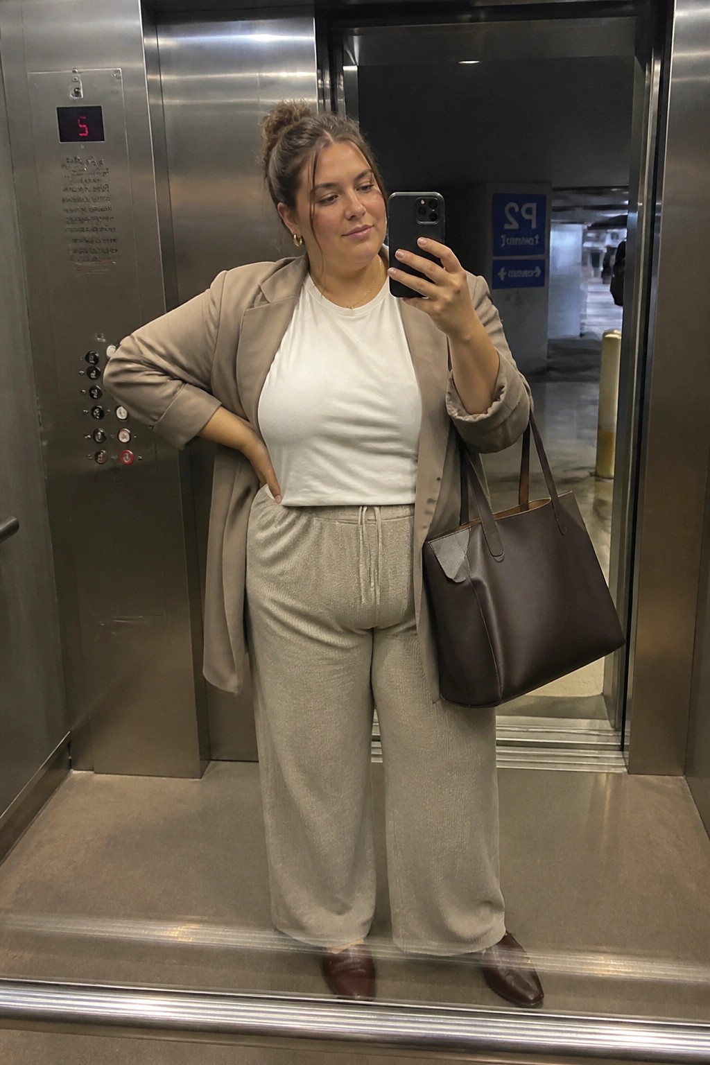 Plus size woman in beige oversized blazer over white t-shirt with light gray wide leg drawstring pants, brown leather tote bag, and brown pointed shoes taking mirror selfie in elevator.