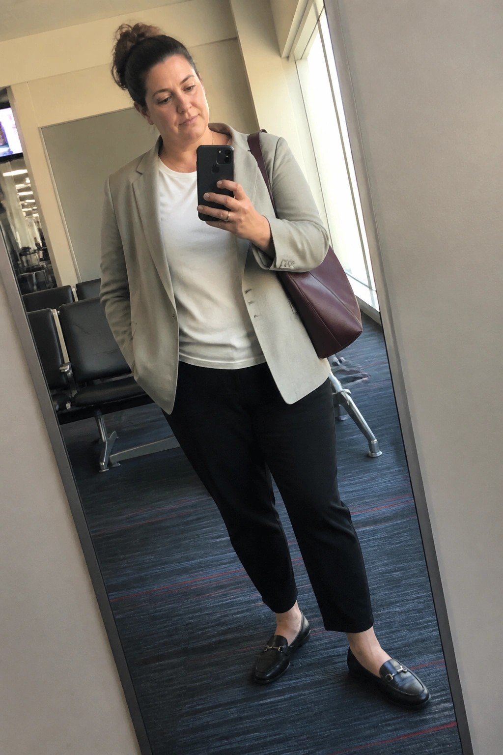 Woman in beige blazer and black pants taking mirror selfie with phone and maroon bag.