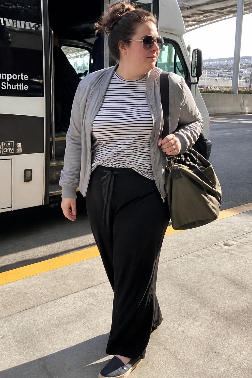 Woman in gray bomber jacket and striped shirt carrying green bag by shuttle bus