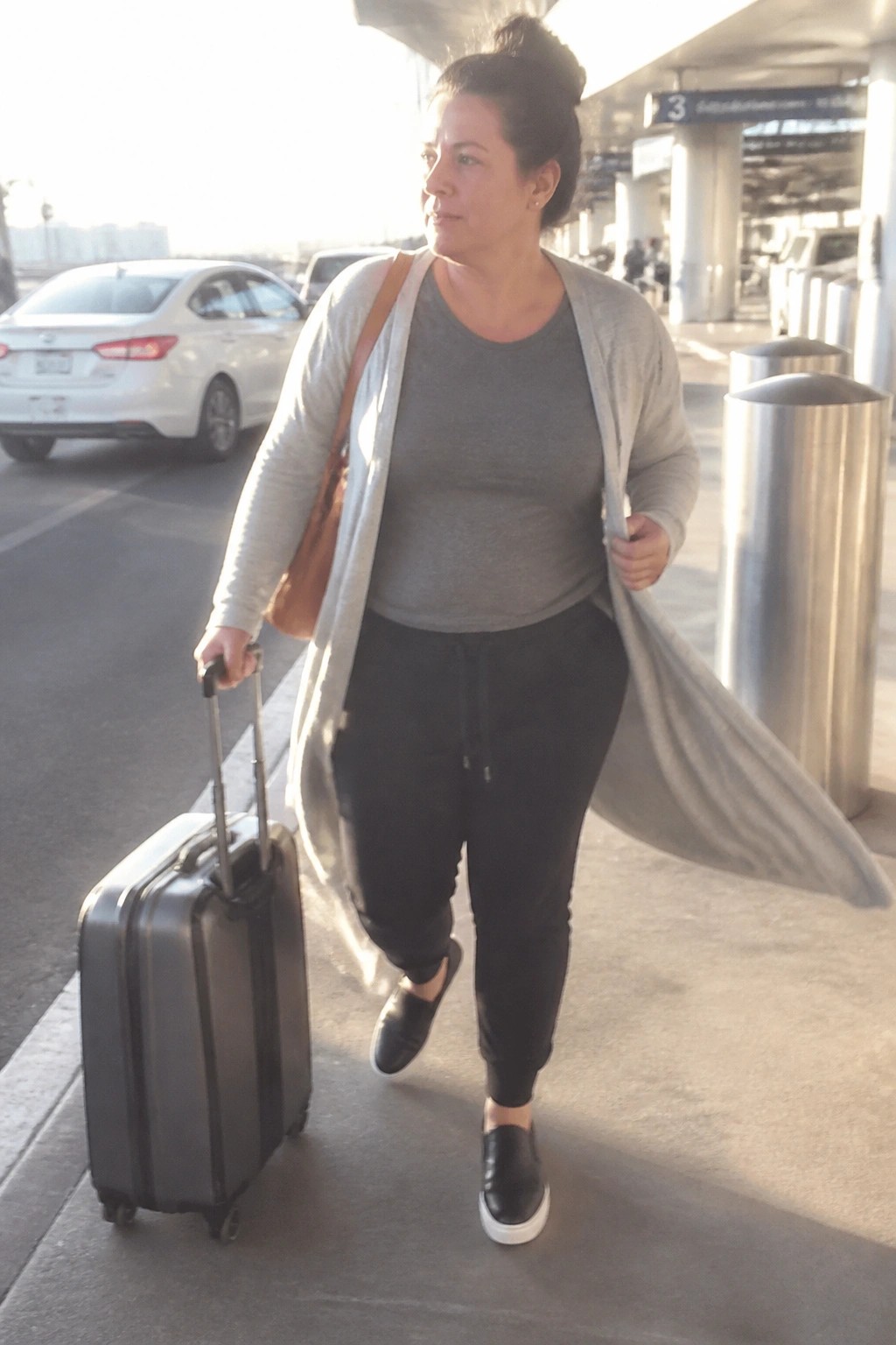 Woman in gray cardigan pulling rolling suitcase at sunny airport terminal