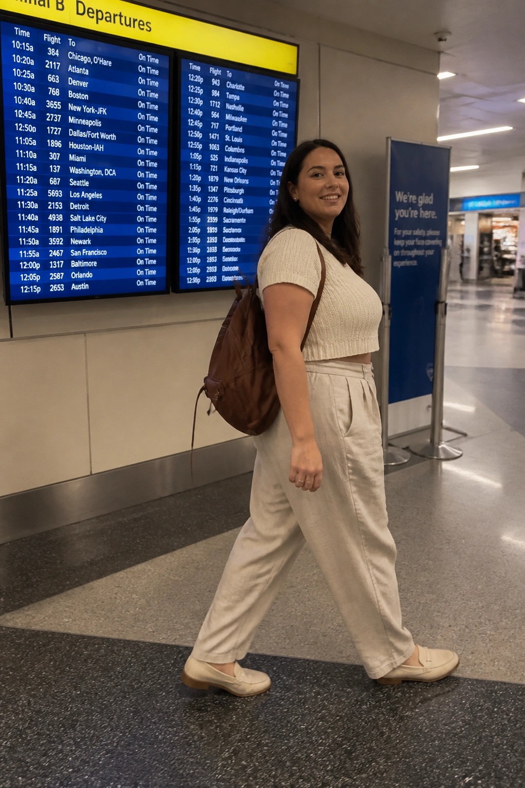 Plus size woman wearing a cream short sleeve knit crop top, beige wide leg trousers, white loafers, and brown leather backpack in an airport terminal next to departure boards.