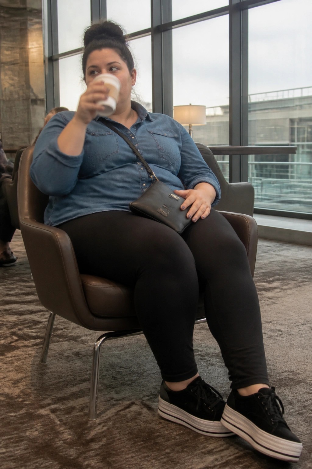 Woman in blue denim shirt drinks coffee in brown armchair with black bag