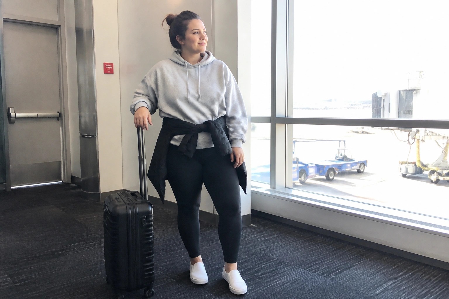 Woman in gray hoodie with black puffer jacket at waist holding suitcase in airport