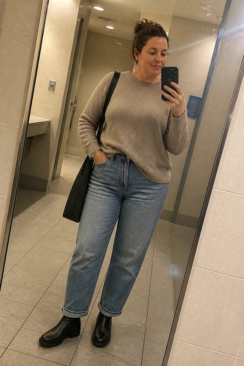 Woman in beige sweater and cuffed blue jeans taking mirror selfie in bathroom