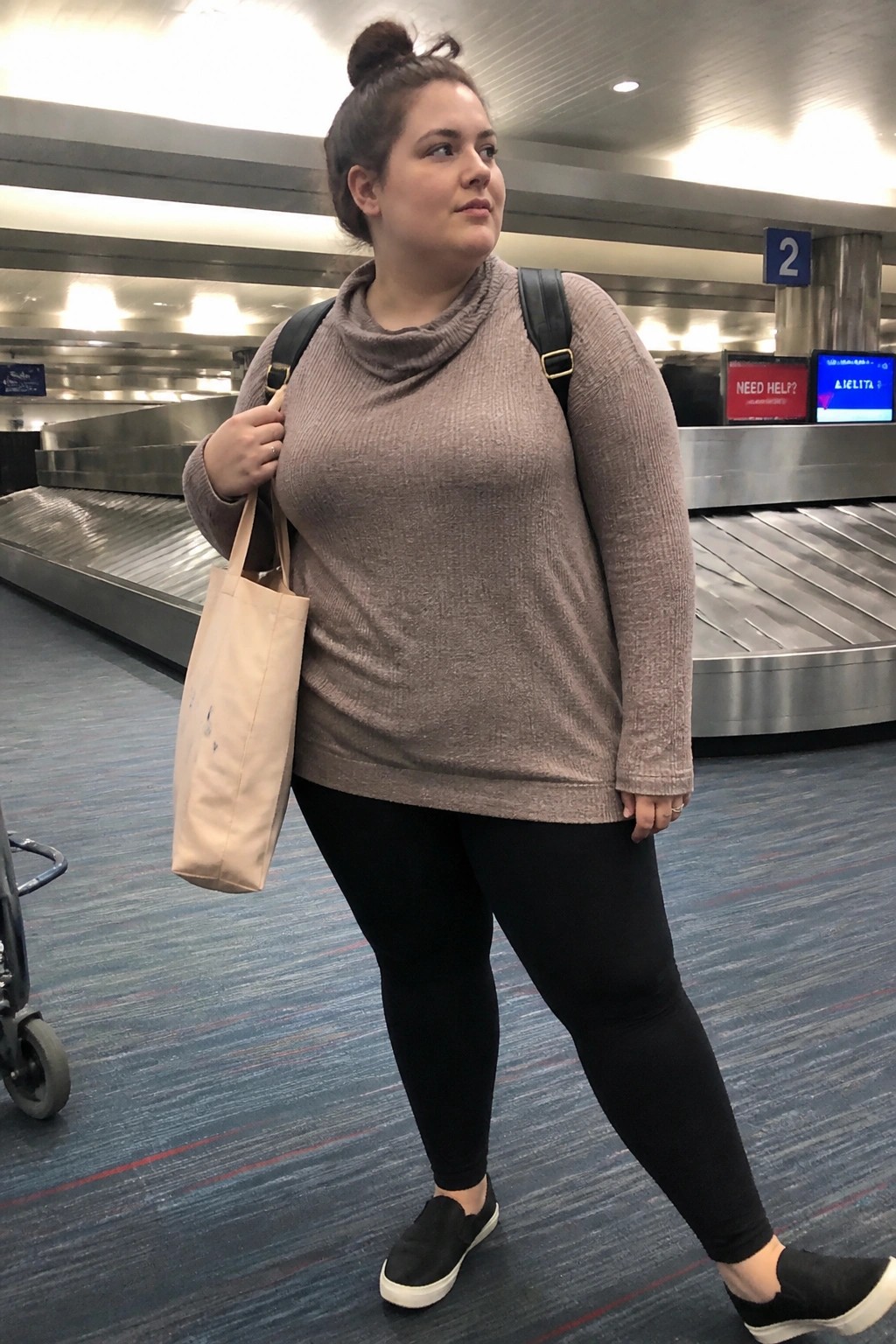 Plus size woman wearing a taupe cowl neck knit sweater with black leggings and black slip on sneakers while holding a beige tote bag near airport baggage claim.