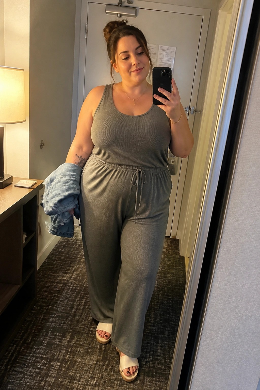 A plus size woman in a gray ribbed tank top and matching wide leg pants holding a light blue denim jacket while wearing white slides in a mirror selfie.