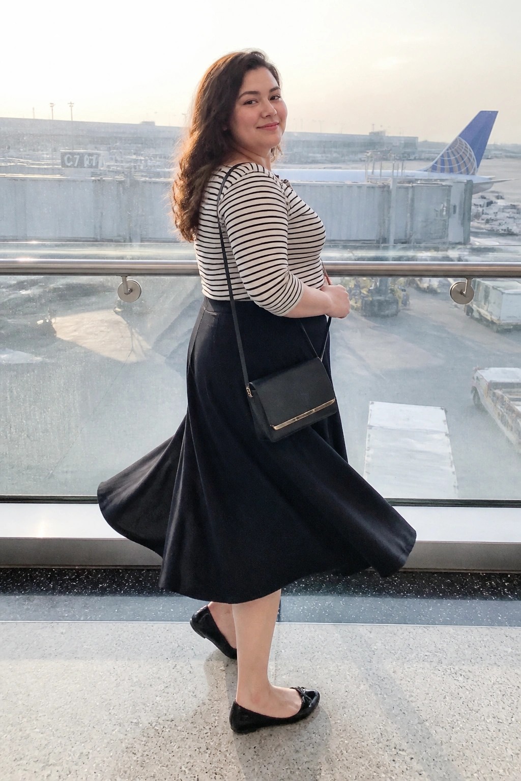 Plus size woman wearing a black and white striped three quarter sleeve top with a flowing black midi skirt, black crossbody bag, and black flat shoes in an airport setting.