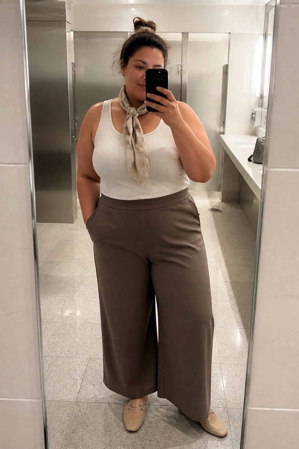 Woman in white tank top, beige scarf, and brown wide-leg pants taking mirror selfie in restroom