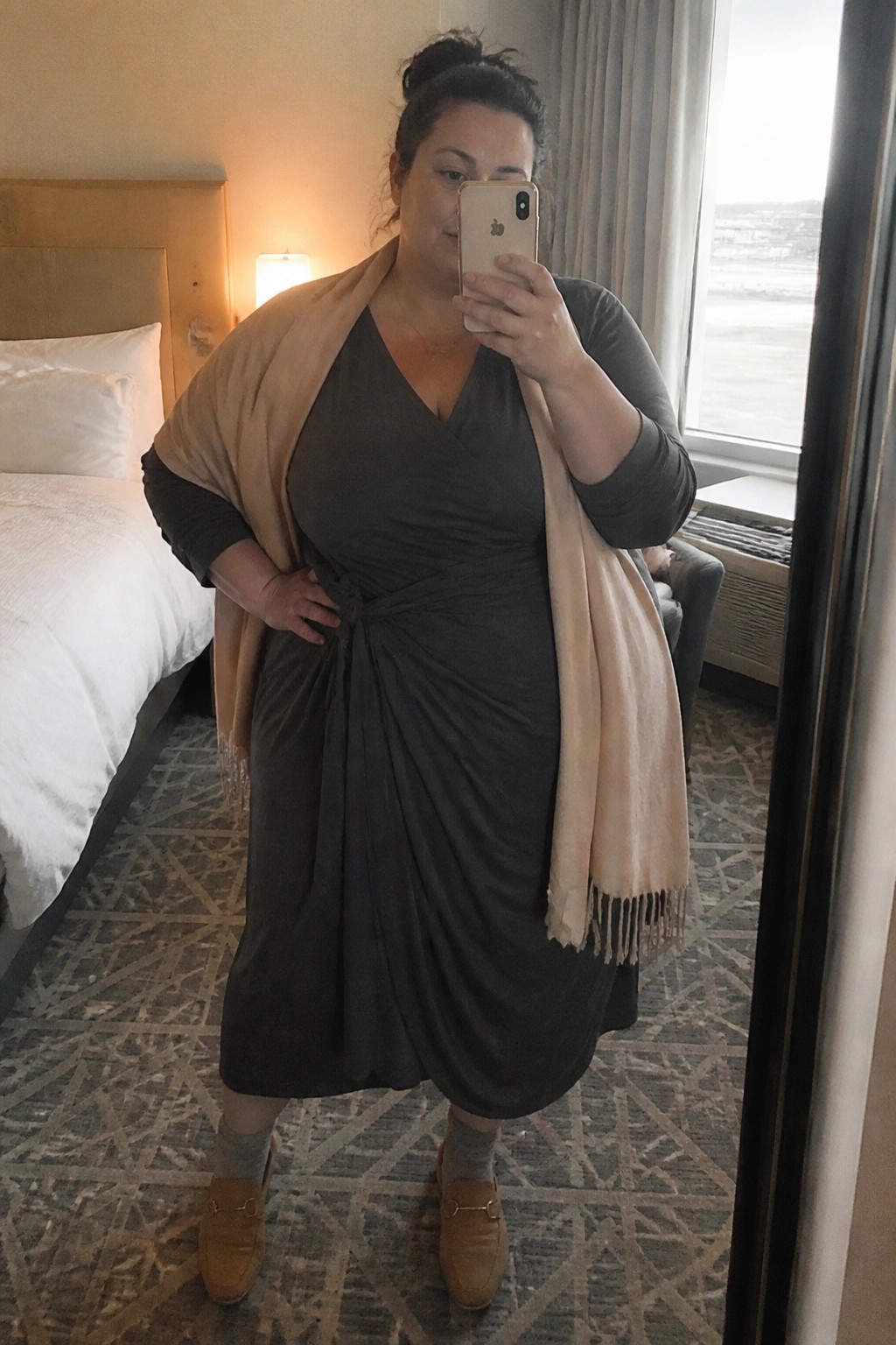 Woman in gray dress and beige shawl taking mirror selfie in hotel room
