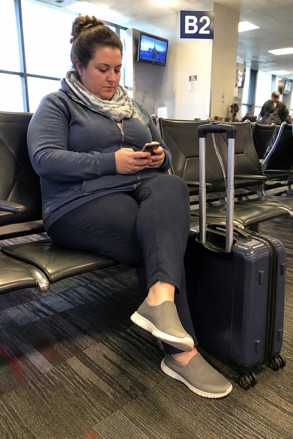 Woman in blue hoodie scrolls phone beside blue suitcase in airport terminal