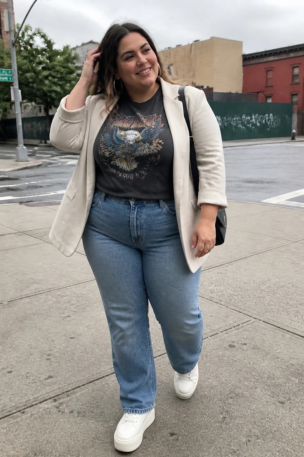 Woman in beige blazer, eagle graphic tee, blue jeans, and white sneakers on city street.
