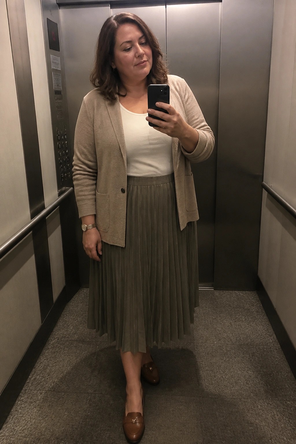 Woman in beige cardigan and olive pleated skirt taking selfie in elevator
