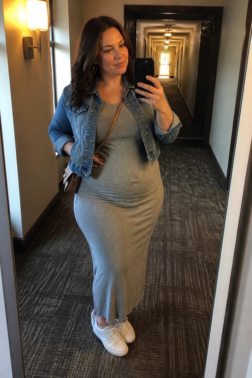 Woman in denim jacket and gray dress taking mirror selfie in hallway