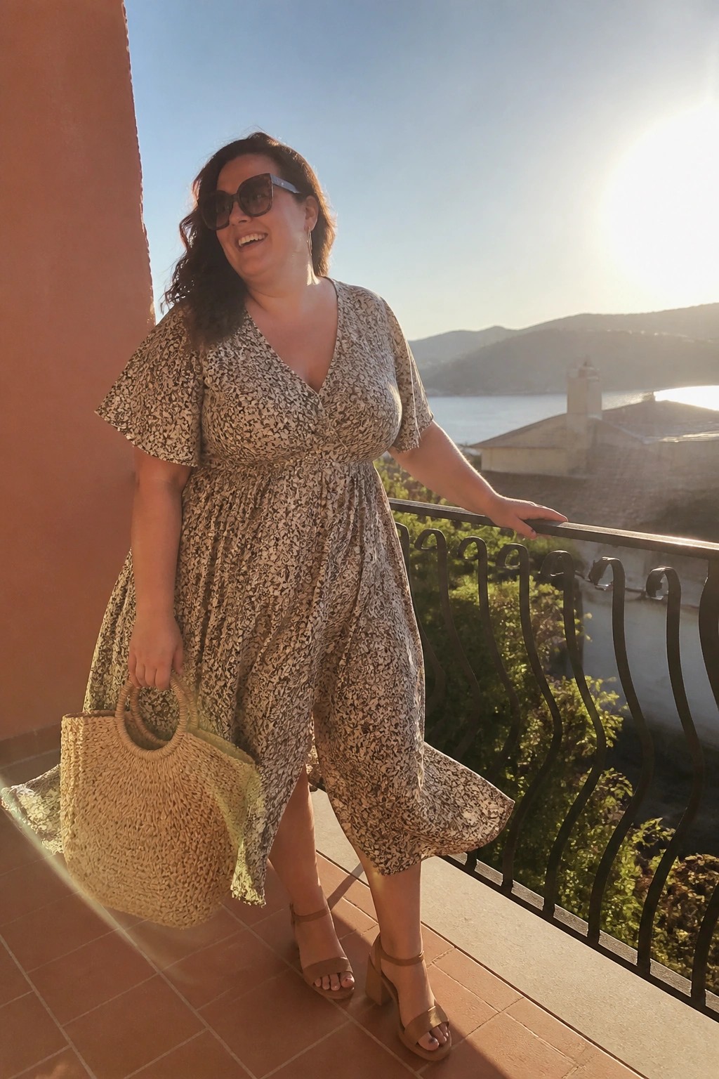 Woman in floral midi dress holding woven tote on sunny sea-view balcony