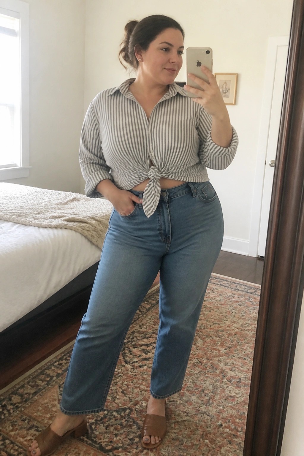 Plus-size woman in striped tied shirt and jeans taking bedroom mirror selfie.
