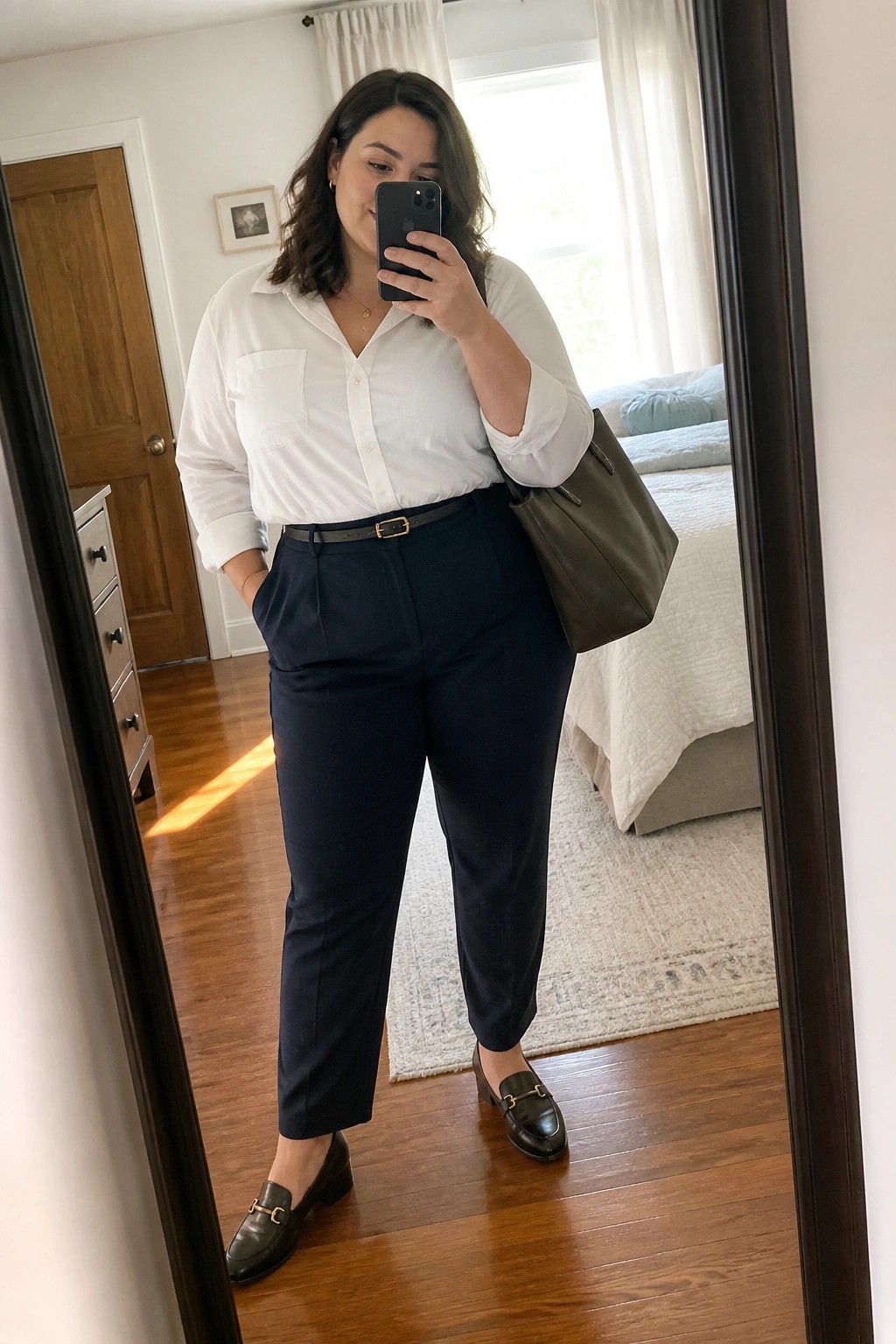 Woman in white shirt and navy pants taking mirror selfie with phone and bag