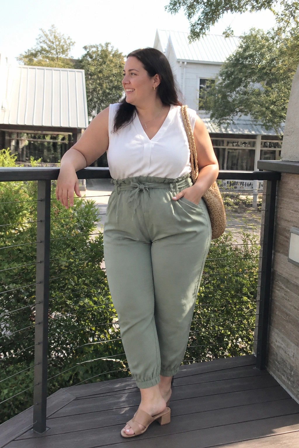 Smiling woman in white top and sage green pants leaning on balcony railing