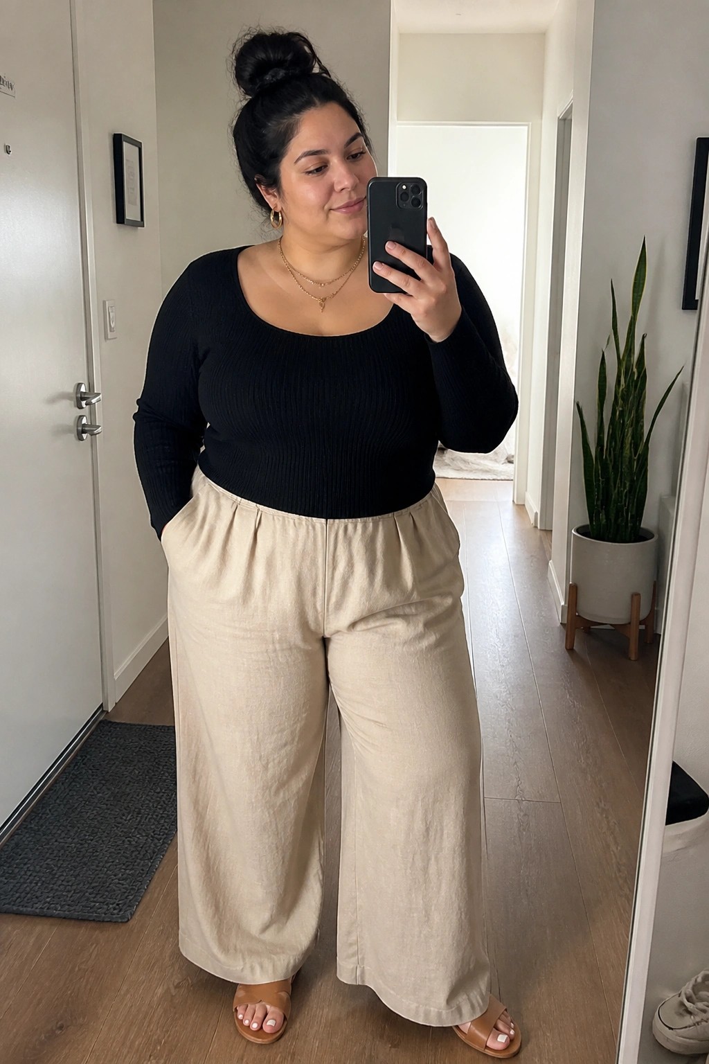 Woman in black top and beige wide-leg pants taking mirror selfie in hallway