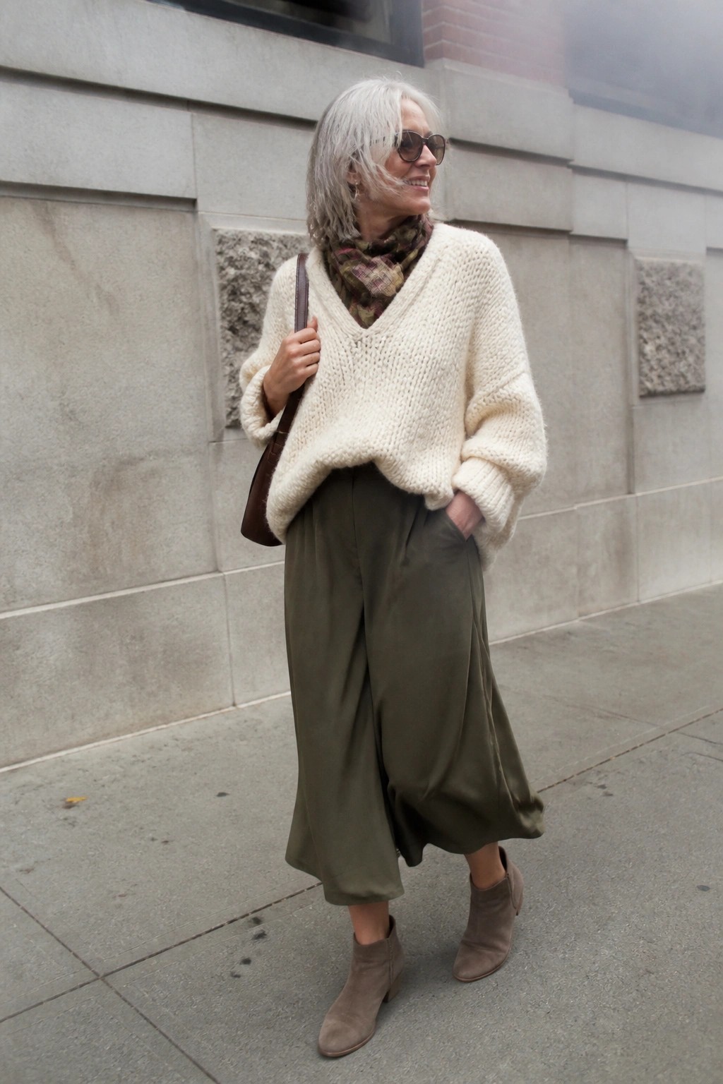 Gray-haired woman in cream knit sweater and olive pants on city sidewalk