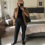 A woman with short gray hair wearing a camel blazer over a navy turtleneck, dark leather pants, and black loafers, holding a brown leather tote bag while taking a mirror selfie in a bedroom.