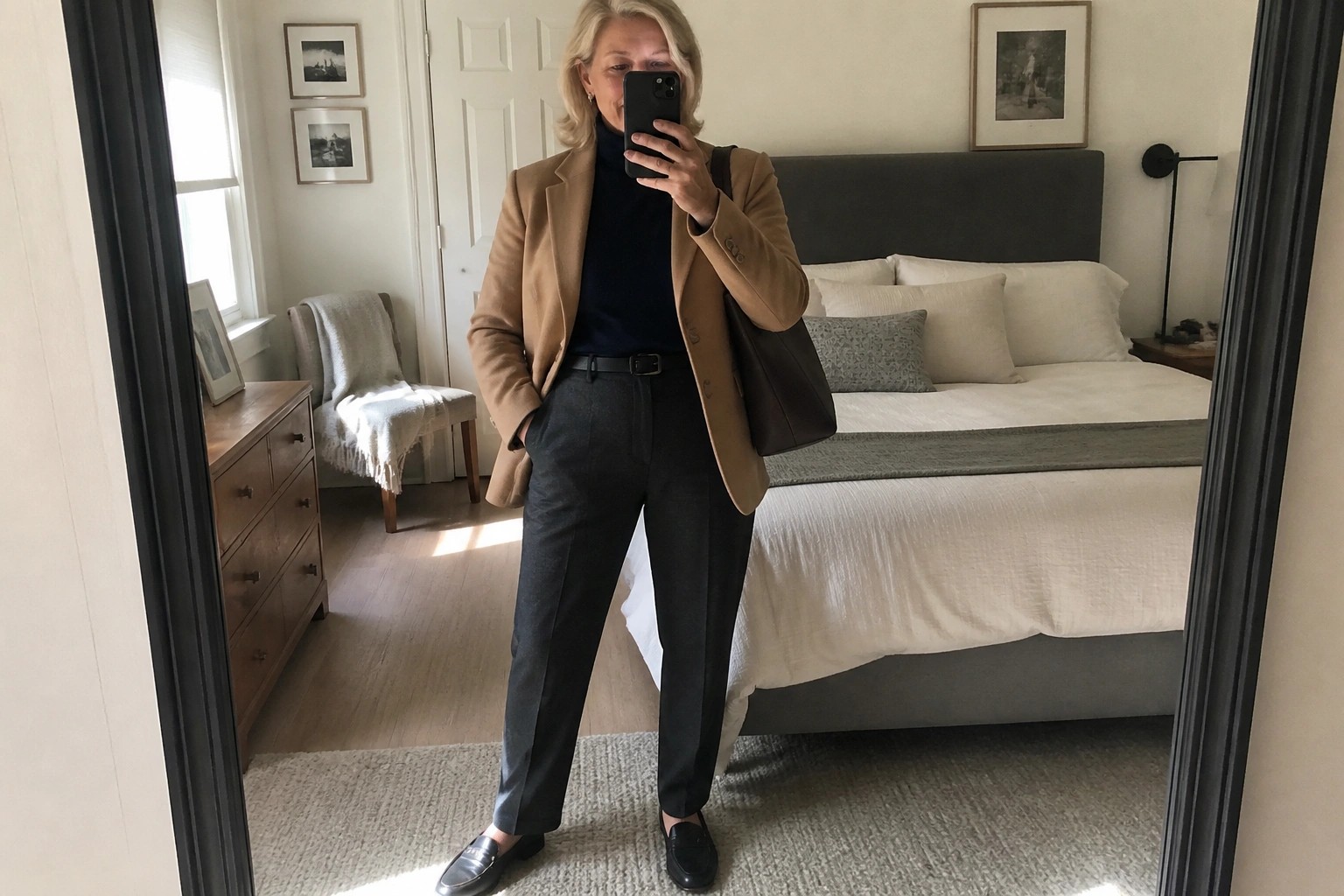 A woman with short gray hair wearing a camel blazer over a navy turtleneck, dark leather pants, and black loafers, holding a brown leather tote bag while taking a mirror selfie in a bedroom.