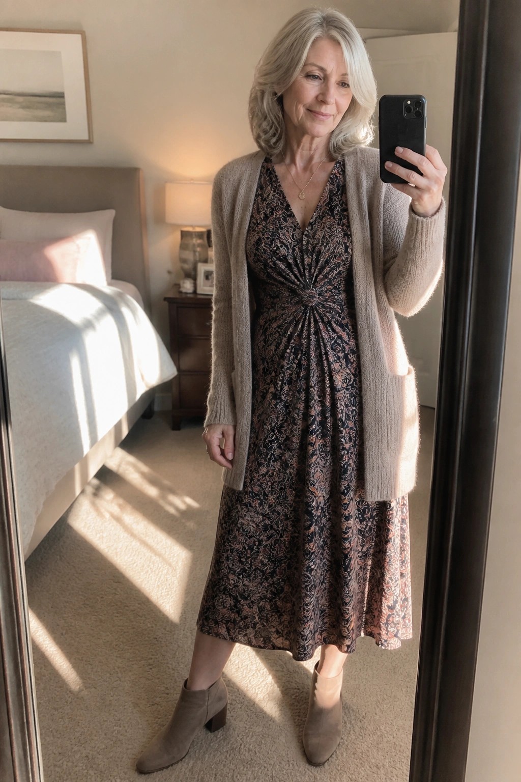 Woman with gray hair wearing a beige open cardigan over a black floral print midi dress with ruched waist and taupe suede ankle boots while holding a phone for a mirror selfie in a bedroom setting