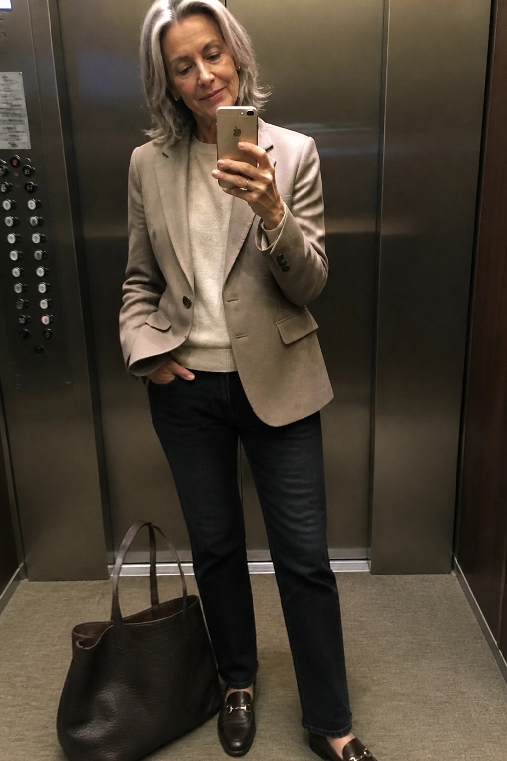Silver-haired woman in beige blazer takes selfie in elevator beside brown tote bag.