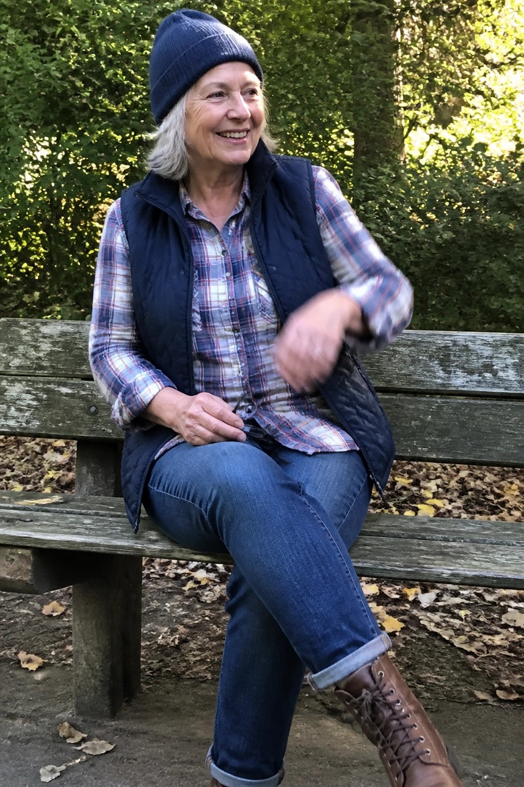 Elderly woman in plaid shirt and blue vest smiles on wooden park bench