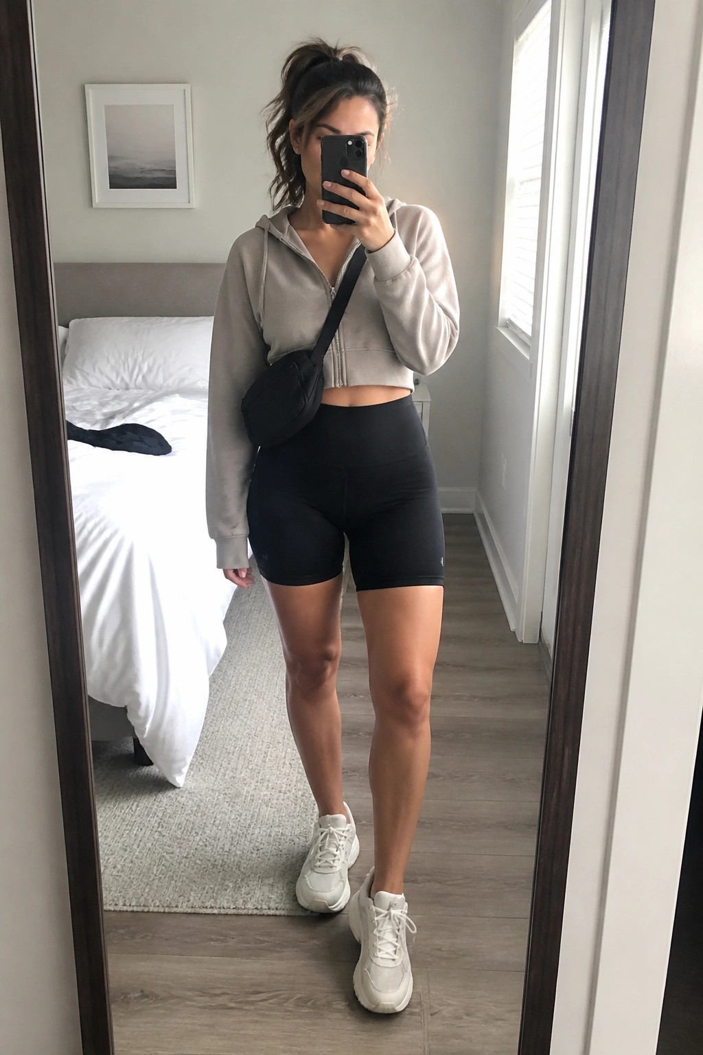 Woman taking a mirror selfie in a cropped gray zip hoodie, black bike shorts, white sneakers, and a small black crossbody bag.