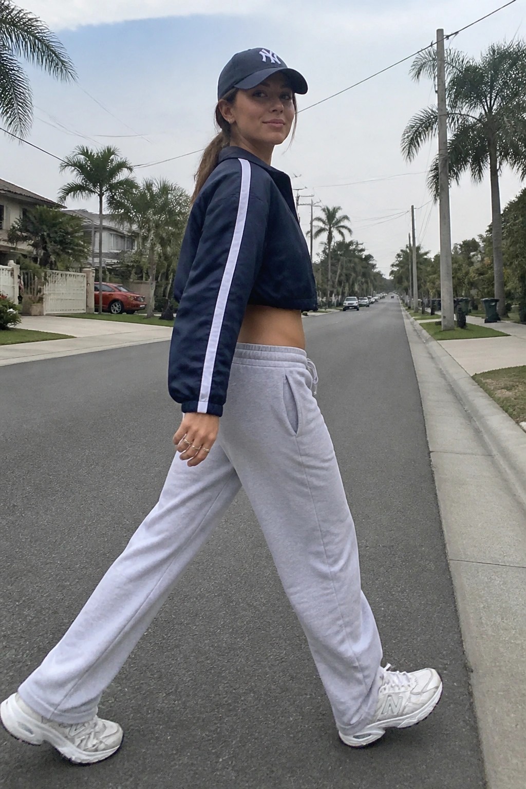 Woman wearing a navy cropped bomber jacket with white sleeve stripes, grey sweatpants, white sneakers, and a navy baseball cap while walking on a street.
