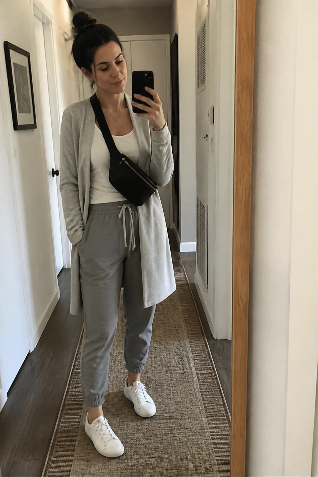 Woman wearing light gray cardigan over white tank top with matching gray joggers, white sneakers, and black crossbody bag in a hallway mirror selfie.