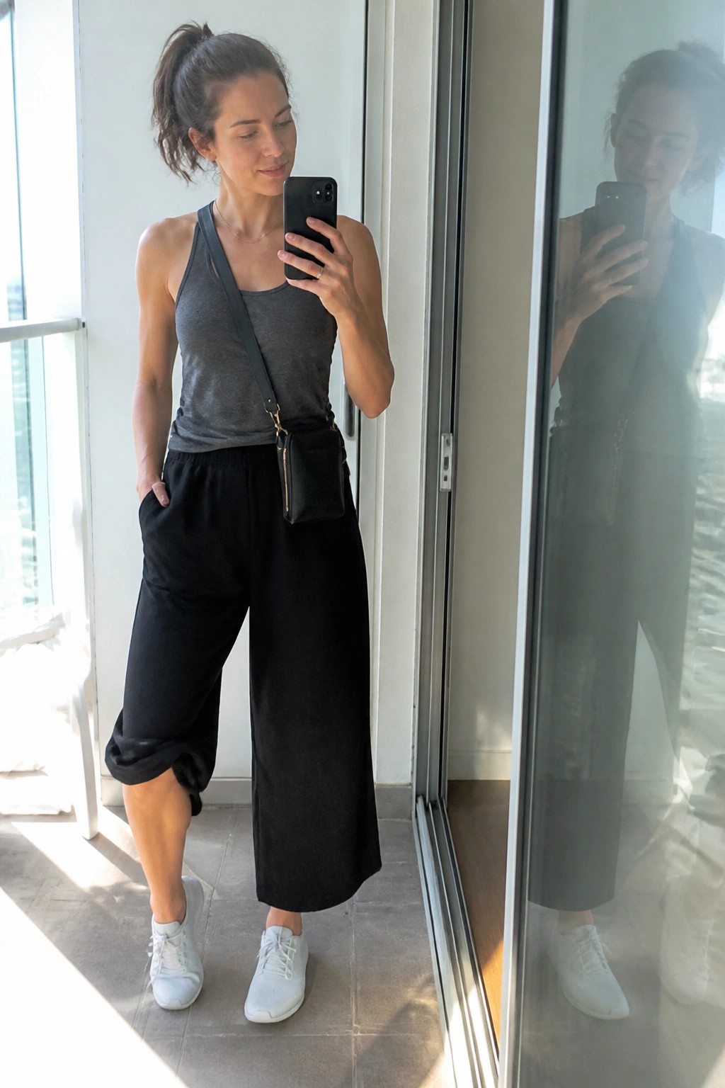 Woman in gray tank top and black pants taking mirror selfie with phone