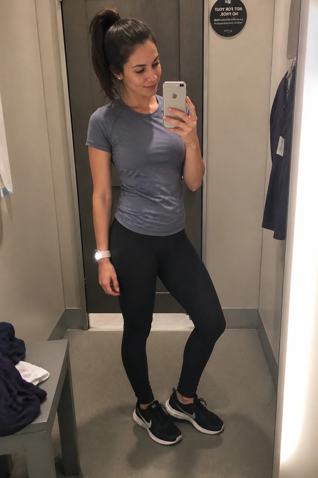 Woman in gray top and black leggings taking mirror selfie in fitting room.