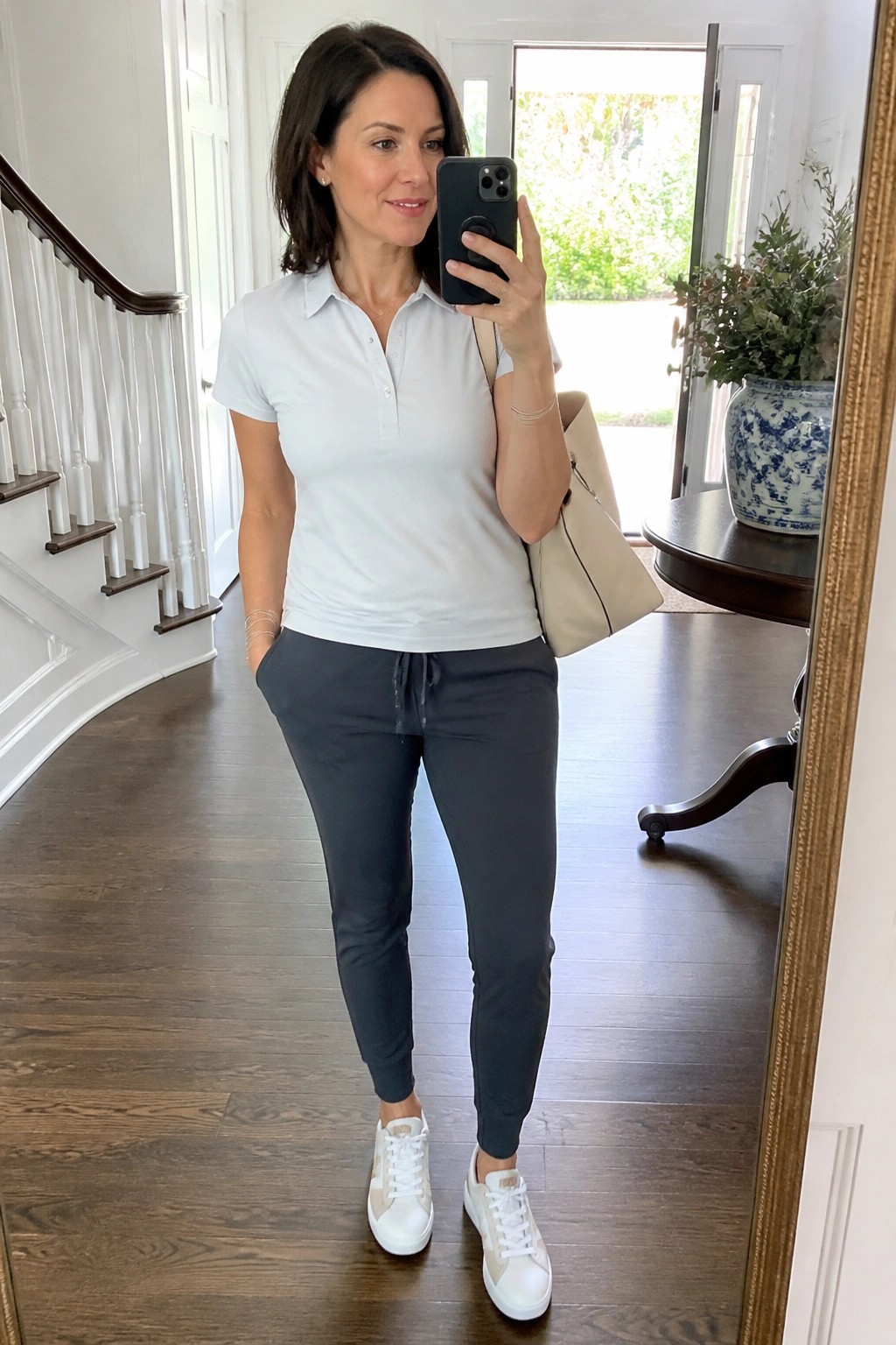 Woman in light blue polo shirt tucked into charcoal jogger pants with white sneakers holding a beige tote and phone for a mirror selfie in an entryway.
