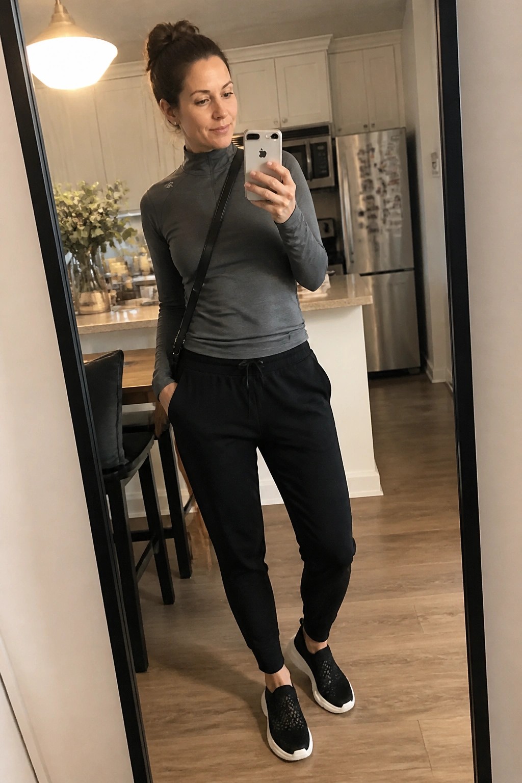 Woman in gray quarter zip top, black jogger pants, and black woven slip on sneakers taking a mirror selfie in a kitchen.