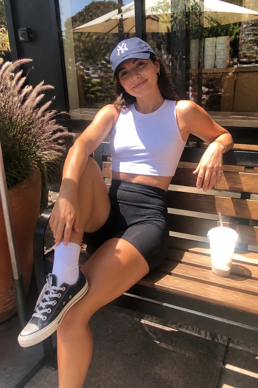 Woman in white crop top, black shorts, blue NY cap on wooden bench with drink.