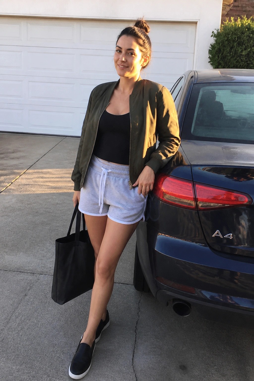 Woman in olive bomber jacket over black tank top, light gray athletic shorts with white trim, and black slip-on sneakers, holding a black tote bag while standing beside a car.
