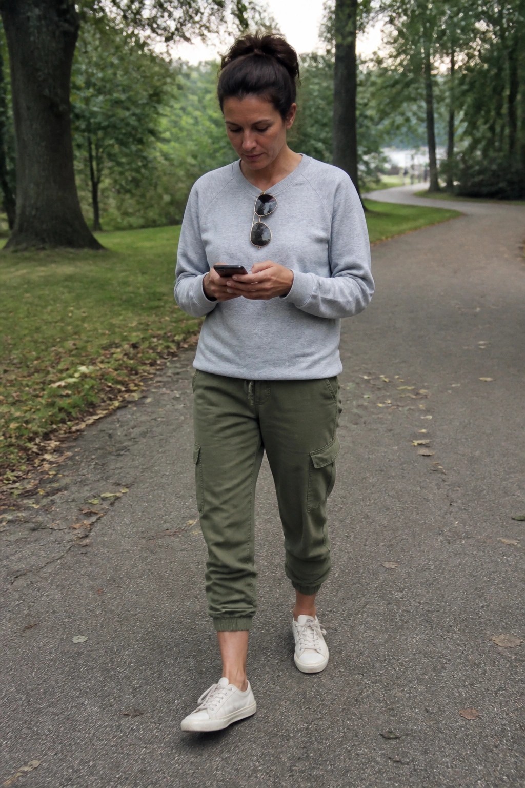 Woman in gray sweatshirt and green cargo pants views smartphone on park path