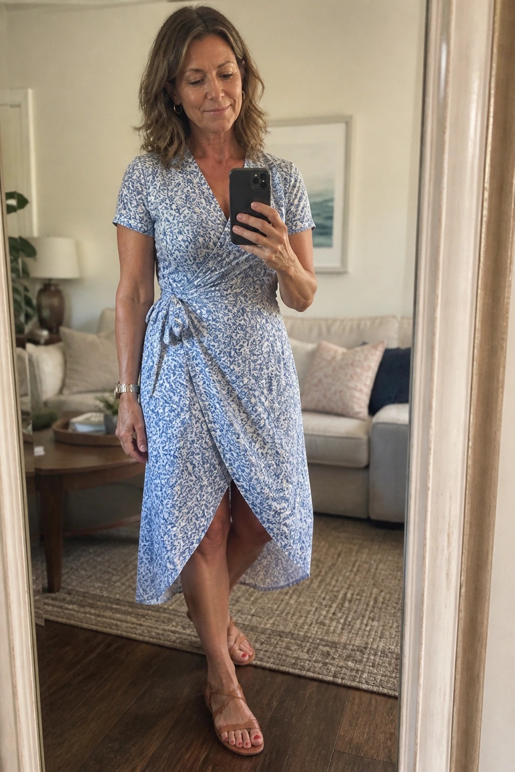 Woman in blue floral wrap dress taking mirror selfie in living room